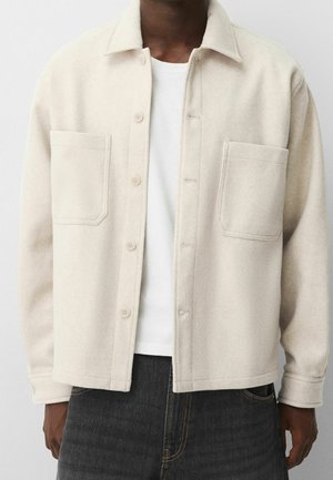 Mężczyzna ubrany w kremową wełnianą koszulę typu overshirt zapinaną na guziki, na białej koszulce, w połączeniu z ciemnoszarymi dżinsami.