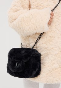 Czarna torba na ramię z sztucznego futra w pikowanym wzorze, z gładkim czarnym łańcuszkiem i prostokątnym kształtem, trzymana w ręce na jasnym tle.