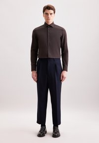 Camicia lunga marrone scuro con colletto classico e chiusura a bottoni, abbinata a pantaloni a pieghe blu navy e scarpe in pelle nera.