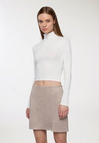 Kobieta z prostymi brązowymi włosami, ubrana w biały, długo rękawowy, krótki top z zamkiem oraz beżową zamszową mini spódniczkę, na tle białym.