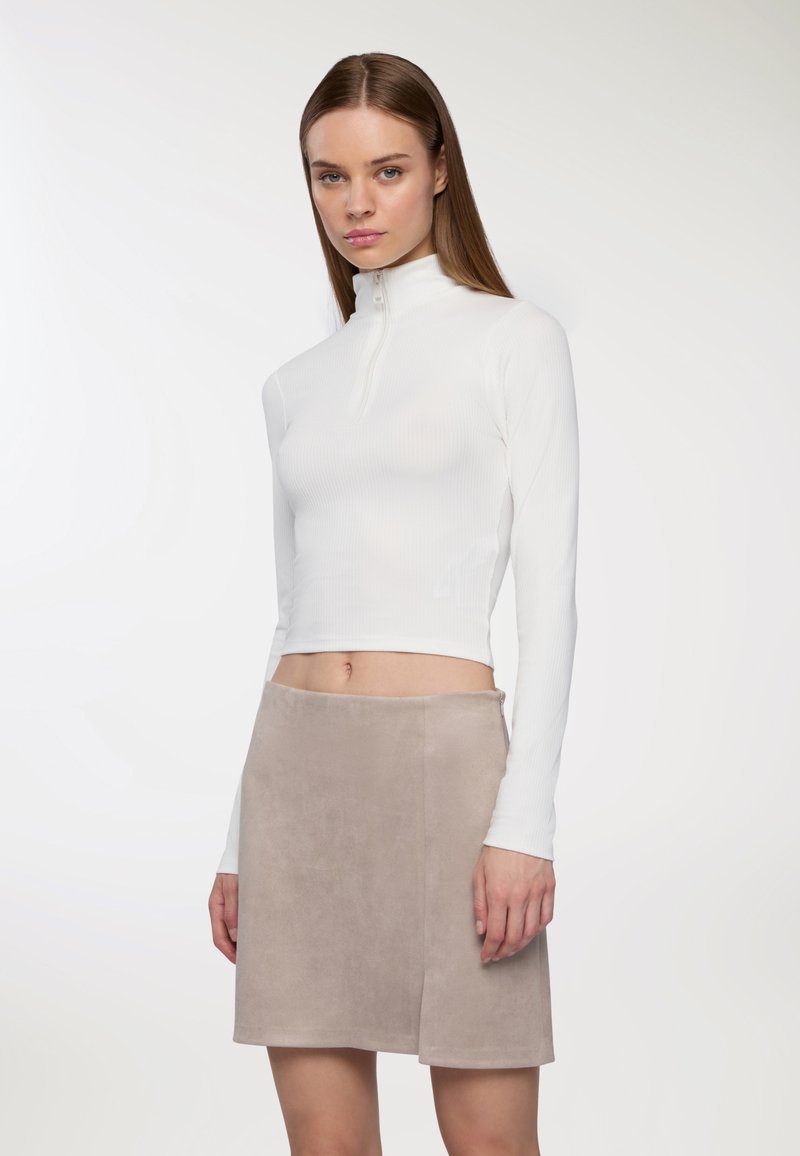 Kobieta z prostymi brązowymi włosami, ubrana w biały, długo rękawowy, krótki top z zamkiem oraz beżową zamszową mini spódniczkę, na tle białym.