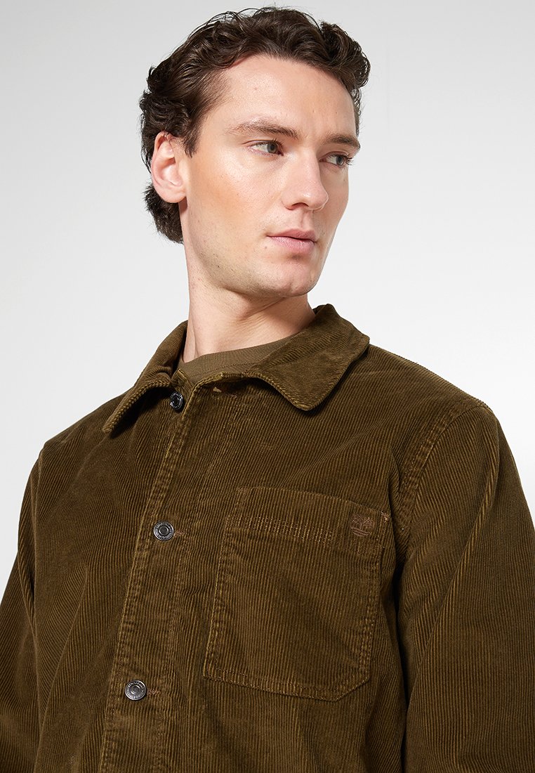 Brązowa kurtka z korduroy z zapięciem na guziki z przodu, kołnierzem i kieszenią na piersi. Posiada pionową ryflowaną teksturę i czarne guziki.