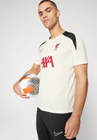 Maglietta da calcio crema con spalle nere, caratterizzata dal logo rosso del Liverpool FC e dal testo "AXA". Il modello tiene in mano un pallone da calcio a fantasia bianco e nero.