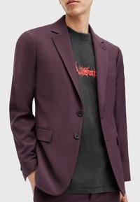 Blazer violet en tissu lisse, à simple boutonnage, avec deux boutons noirs et une poche poitrine, porté sur un t-shirt graphique noir.