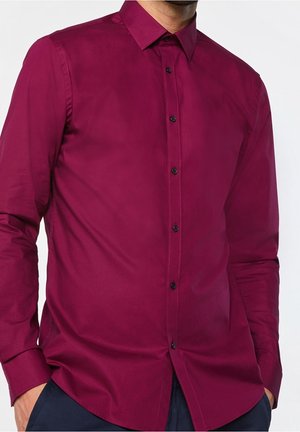 Man draagt een bordeauxrode blouse met lange mouwen en zwarte knopen, handen in de zakken, tegen een effen witte achtergrond.