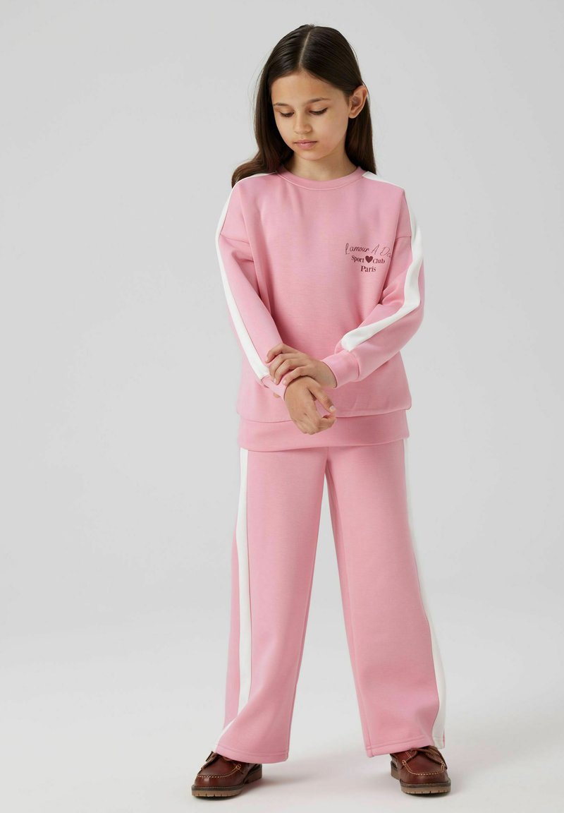Conjunto de sudadera y pantalones en rosa claro, con franjas laterales blancas y una pequeña impresión del logo. Tela suave con un corte relajado.