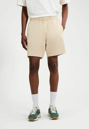 Mann, der beige Shorts mit elastischem Bund, weiße Socken, grüne Turnschuhe und ein weißes T-Shirt trägt, steht vor einem schlichten weißen Hintergrund.