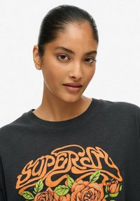 T-shirt nera con testo arancione "Superdry" e grafica floreale di rose. Maniche corte, colletto a giro, tessuto leggero, vestibilità casual.