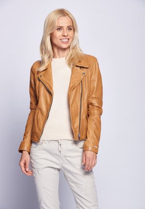 Brun lädermotorcykeljacka med en asymmetrisk dragkedja, standardkrage och figurnära design. Bärs över en ljusbeige tröja och ljusa byxor.