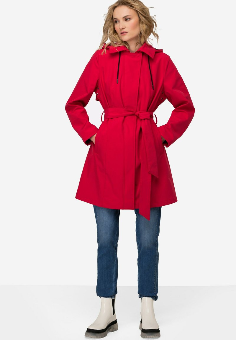 Červený voděodolný trench coat s kapucí na šňůrku, opaskem v pase, bočními kapsami a volným střihem, kombinovaný s modrými džíny a bílými kotníkovými botami.