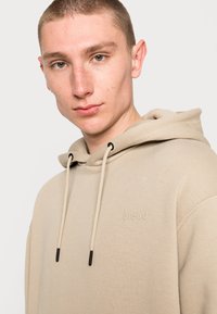 Beige hoodie made from soft fabric, featuring a drawstring hood, black accent tips, and a subtle embossed logo on the chest.