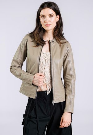 Giovane donna con giacca di pelle beige sopra un top in crochet crema e pantaloni neri, rivolta in avanti con una mano che afferra la cerniera della giacca.