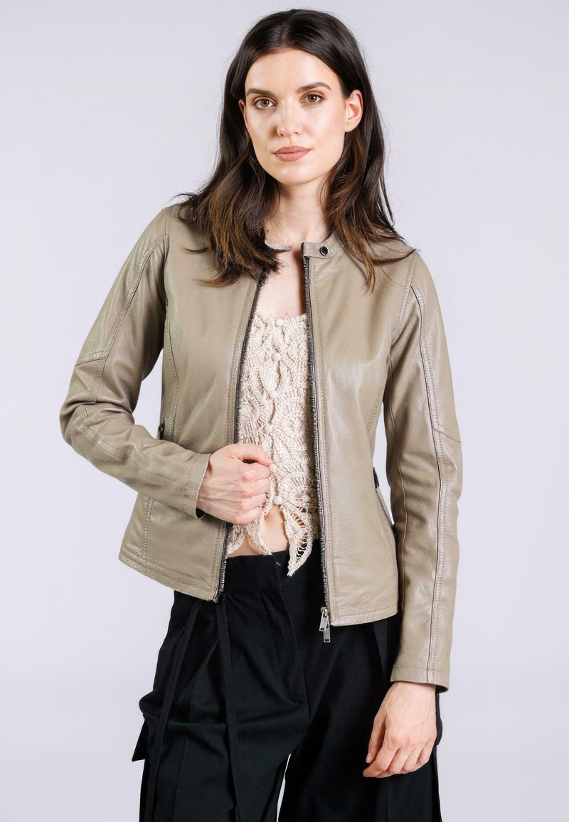 Jonge vrouw draagt een beige leren jas over een crèmekleurige gehaakte top en zwarte broek, kijkt naar voren met één hand die de rits van de jas vasthoudt.
