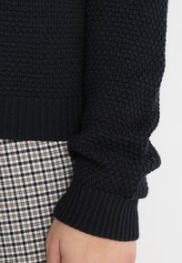 Marinblå stickad tröja med texturerat mönster och ribbade muddar, ihop med rutigt tyg som syns nedanför. Fokusera på material och detaljer.