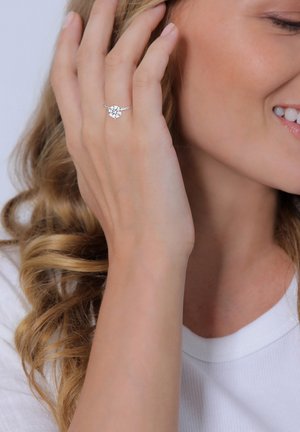 Silver ring with a floral design featuring a central clear stone, worn on a hand with light skin and wavy hair, against a plain background.