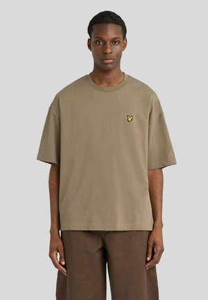 Jonge man draagt een loszittend beige T-shirt met een klein geel vogel-logo op de linkerborst en bruine broek, staand voor een effen achtergrond.