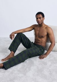 Grønne og marineblå ternede pyjamasbukser med elastik i taljen, der har en sidelomme og en afslappet pasform, båret af en person uden trøje.