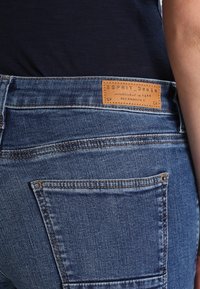 Jeans de denim azul con un acabado texturizado, que presentan un parche de cuero marrón etiquetado como "ESPRIT Denim" en la cinturilla y en el bolsillo trasero visible.