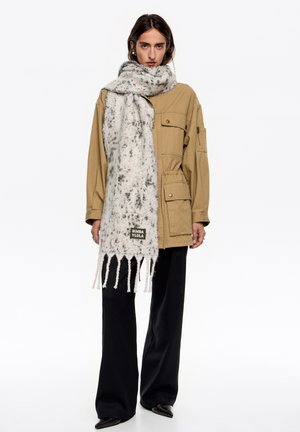 Wełniany szalik w kolorze białym z czarnymi plamkami, z frędzlami na krawędziach oraz metką. Miękka struktura, prostokątny kształt i odpowiednia długość do owijania.