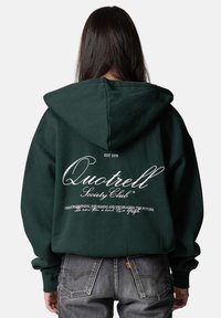 Grüne Kapuzenjacke mit einem großen, weißen Drucklogo auf der Rückseite, das kursiven Text zeigt, und einer Kordelzugkapuze. Weicher Stoff und lockere Passform.
