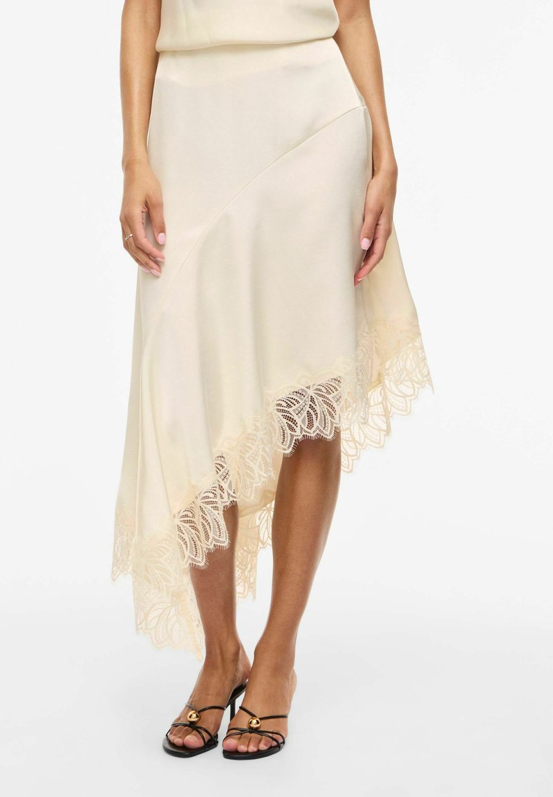 Femme portant une jupe asymétrique crème avec un ourlet en dentelle et des sandales à talons noires à brides, debout sur fond blanc.