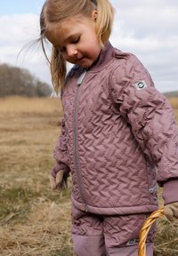 Giacca trapuntata rosa chiaro con colletto e polsini a coste, cerniera argento e toppa con logo; abbinata a pantaloni coordinati e guanti marroni.