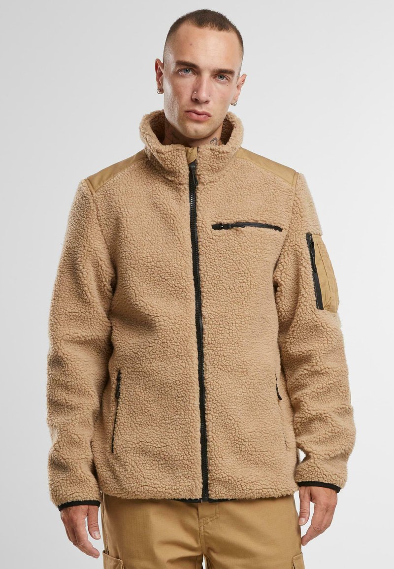 Giacca in pile beige con colletto alto, chiusura con zip nera, tasche laterali e inserti in pelle sulle spalle. Materiale testurizzato e accogliente.