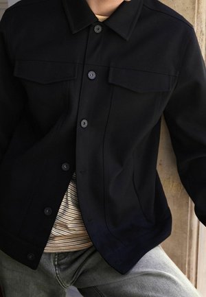 Czarna kurtka z gładkiej tkaniny z kołnierzem, wyposażona w dwie kieszenie na piersi i widoczne zapięcia na guziki. Pod spodem koszula w paski.