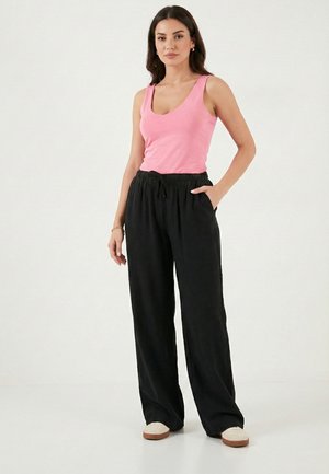 Mujer de pie con una blusa sin mangas rosa, pantalones anchos negros con cordón, zapatos beige y una pulsera, sobre un fondo blanco.