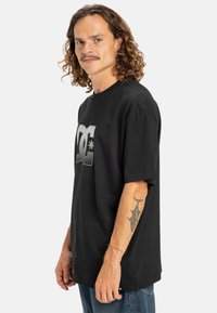 Black cotton t-shirt featuring a large, textured DC logo in gray. The shirt has a relaxed fit and short sleeves.