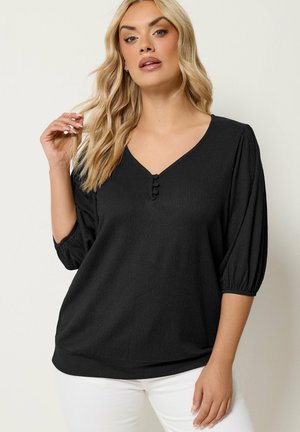 Woman with blonde hair wearing black V-neck blouse with three buttons and elbow-length sleeves, paired with white pants, posing against white wall.