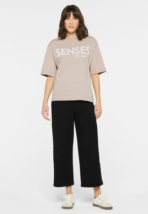 Light beige oversized t-shirt with "SENSES .THE LABEL" printed in white. Paired with black wide-leg pants and light sneakers. Minimalist style.