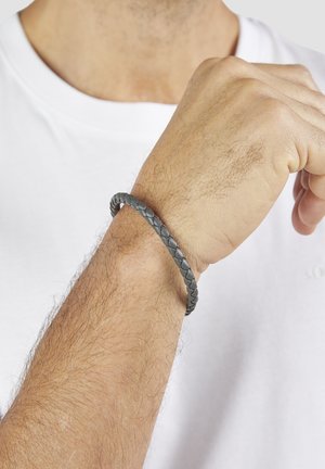 Gray braided leather bracelet on wrist, round shape, smooth texture, subtle sheen, worn over a white shirt.
