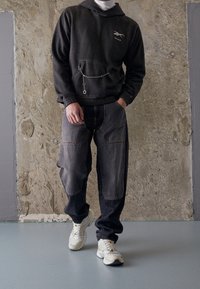 Felpa grigia con tasca frontale e dettaglio in catena, abbinata a pantaloni in denim scuro con più tasche e sneakers chunky di colore chiaro.