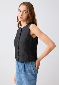 Femme portant un haut noir sans manches à boutons et un jean bleu, debout avec une main sur la hanche devant un fond uni.