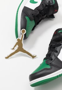 Paire de baskets Air Jordan noires et vertes avec lacets noirs, semelles intermédiaires blanches et une étiquette logo Jumpman dorée à proximité sur une surface blanche.