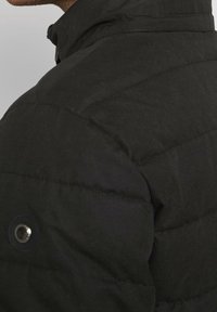 Detailní záběr na rameno černé prošívané bundy s prošívaným stehem a zaobleným černým knoflíkem na světle šedém pozadí.