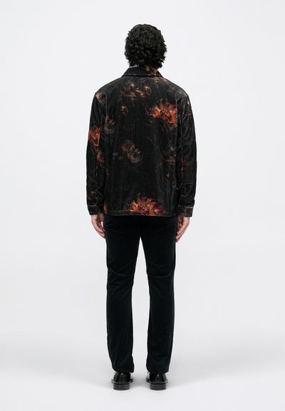 Jachetă din catifea neagră cu un model floral multicolor pe spate, asortată cu pantaloni din catifea întunecată și pantofi negri, văzută din spate.