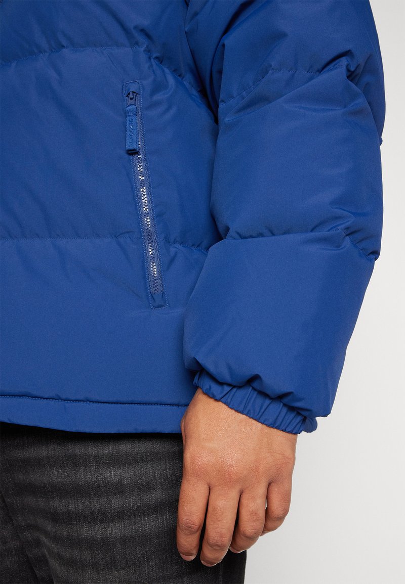 Giacca a vento blu con polsini elasticizzati e tasca con zip. Finitura liscia e opaca con sezioni trapuntate per l'isolamento.