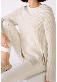 Personne portant un pull en maille côtelée crème avec une fermeture éclair latérale et un pantalon écru, assise avec un genou relevé et la main posée dessus.