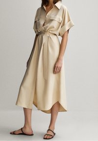 Beige satiininen mekko, jossa on lyhyet hihat ja vyötärösolmu; edessä napitus, kaulus ja epäsymmetrinen helma. Yhdistetty mustiin sandaaleihin.