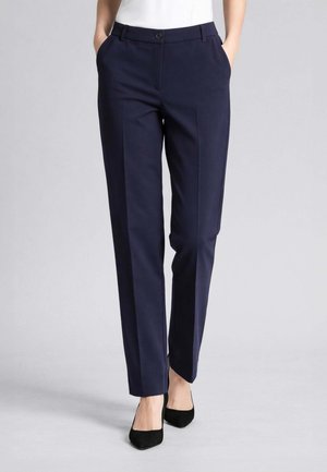 Donna che indossa pantaloni sartoriali blu scuro a gamba dritta con piega frontale e tasche laterali, abbinati a décolleté nere a punta e top bianco.