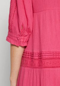 Vestido rosa com tecido em camadas, padrão texturizado e acabamento decorativo. Mangas bufantes com fecho de botão e detalhe aberto. Acabamento suave.
