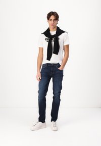 Tricou alb cu logo-ul negru "GUESS", blugi bleumarin, pulover negru aruncat pe umeri și adidași albi. Material neted și croială clasică.
