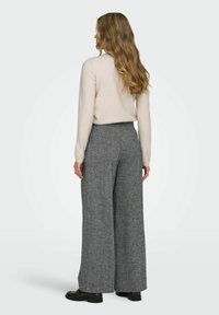 Moteris su ilgais banguotais plaukais dėvi kreminę megztinį, aukšto liemens plačius pilkus kelnes ir tamsius loafers, stovėdama atsukusi nugarą prieš vientisą foną.