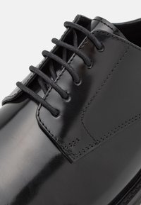 Schwarze Lederdressschuhe mit glänzender Oberfläche, sechs Schnürsenkellochern und verstärkten Nähten entlang der Kante für zusätzliche Haltbarkeit.