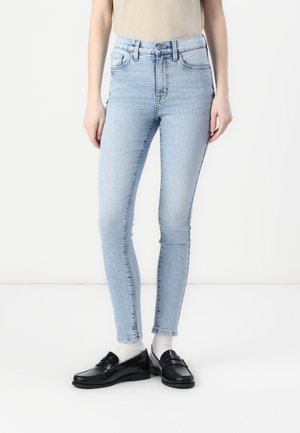 Helle blaue, hochtaillierte Skinny-Jeans mit glatter Textur, die über fünf Taschen und Gürtelschlaufen verfügt, kombiniert mit schwarzen Slippers.