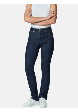 Person iført mørkeblå højtaljede skinny jeans, hvid tanktop og hvide sneakers, stående med armene krydset.