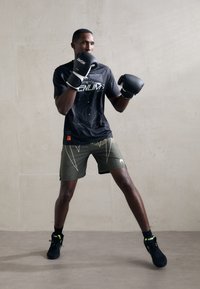 Camisa negra de manga corta con estampado gráfico, pantalones cortos verdes con acentos amarillos, guantes de boxeo negros y zapatillas negras con acentos neón.