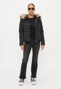Casaco acolchoado preto com capuz de pele sintética, fecho de zip, usado sobre uma blusa preta com pontos prateados, combinado com jeans escuros de boca de sino e ténis.